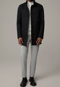 Svart, knapplång kappa med hög krage; bärs över en vit topp och grå byxor. Modellen har på sig svarta skor. Minimalistisk design, slät textur.