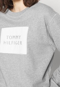 Grey sweatshirt with a white rectangular patch featuring "TOMMY HILFIGER" in embossed lettering. Soft fabric, crew neckline, and ribbed cuffs.