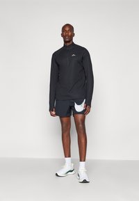 Svart langermet jakke med glidelås, lett stoff, pyntet med mørke shorts med en flekket lomme. Modellen står i sportssko og sokker.