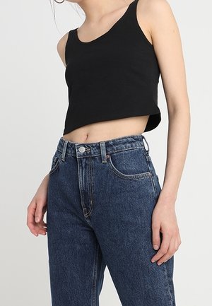 Femme portant un haut court noir sans manches et un jean en denim bleu taille haute sur un fond uni.