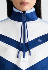 Pullover Lacoste blu e bianco con colletto alto, chiusura con zip, motivo a zigzag e logo ricamato. Tessuto liscio e sportivo.