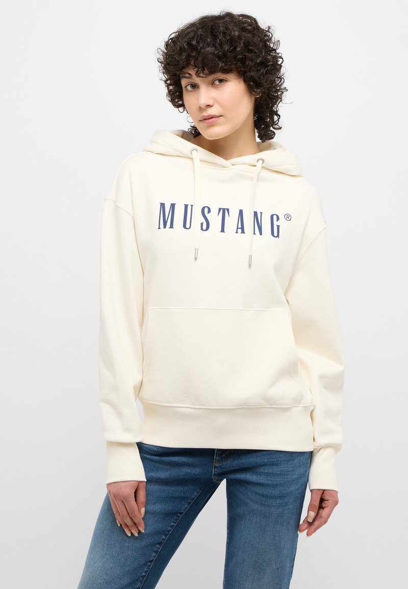 Sudadera color crema con bolsillo canguro frontal, que presenta un prominente logo azul de "MUSTANG" y capucha con cordón. Combinada con jeans azules.