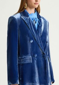 Blazer doppiopetto in velluto blu con rever a lancia, due tasche frontali e bottoni rotondi, che mostra una lucentezza morbida e una texture liscia.