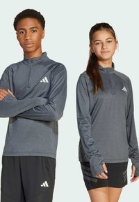 Tmavošedá sportovní trička s dlouhým rukávem, čtvrtinovým zipem a logem. Oba nosí černé kraťasy s designem na boku.