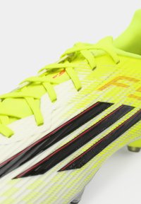 Λαχανί ποδοσφαιρικό παπούτσι με υφή στην επιφάνεια, με μαύρες και κόκκινες λεπτομέρειες, έντονα κορδόνια και καμπυλωτή σχεδίαση για βελτιωμένη απόδοση.