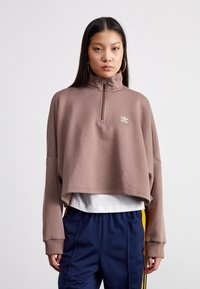 Sweatshirt castanho cropped com gola alta e fecho frontal. Apresenta mangas longas e um logótipo no peito. Emparelhado com calças azuis-marinho com riscas amarelas.