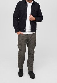 Svart knäppjacka över en vit t-shirt, ihop med olivgröna cargobyxor med flera fickor, och svarta snörade stövlar.