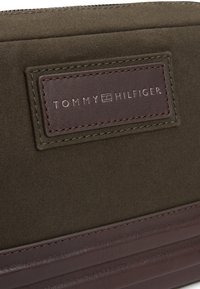 Olivengrønn stofflommebok med brunt lærdetalj med preget "TOMMY HILFIGER"-logo og kontraststing. Glidelås lukking øverst.