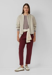 Vrouw in een beige zip-up jack, gestreept langemouwen shirt, bordeauxrode broek, beige trui om haar taille geknoopt en beige sneakers staand tegen een grijze muur.