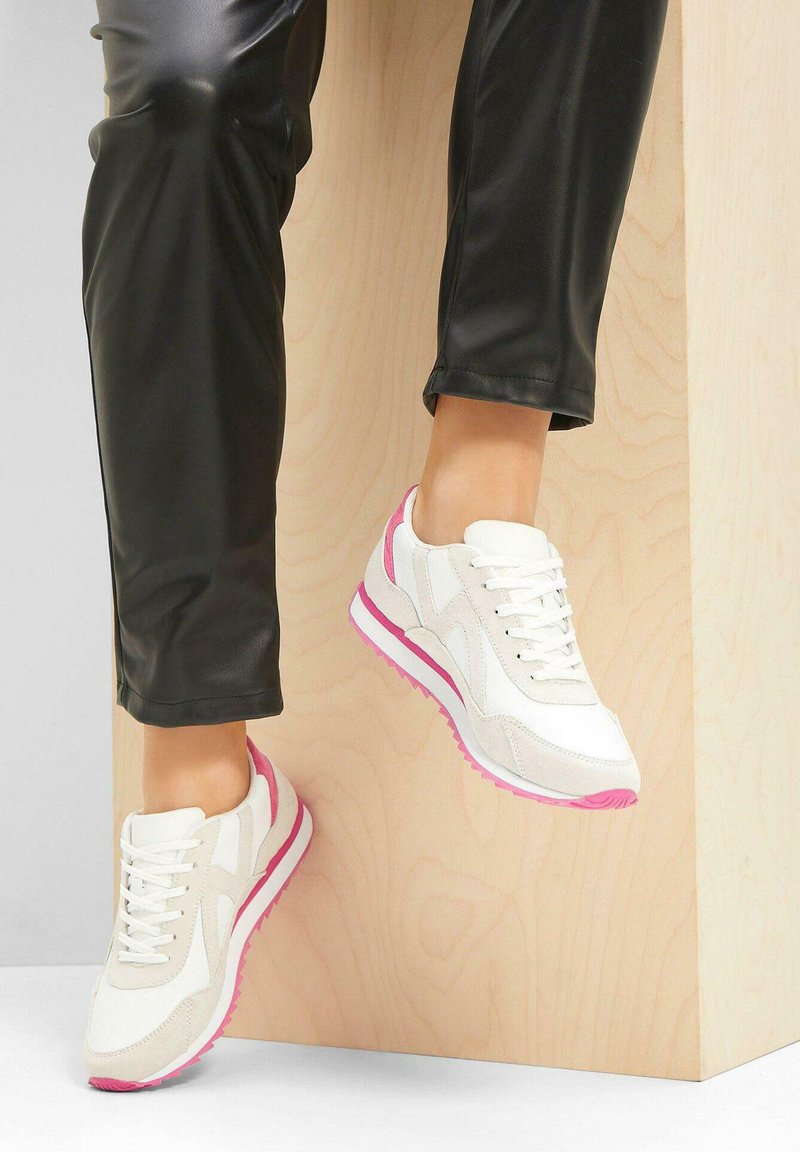 Sneaker bianchi e beige chiaro con accenti rosa, con una texture liscia, chiusura con lacci e un design retrò, indossati con pantaloni neri.