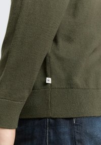 Donkergroene gebreide trui met geribbelde zoom en manchetten. Heeft een klein merketiket aan de zijkant, close-up die de textuur en pasvorm laat zien.