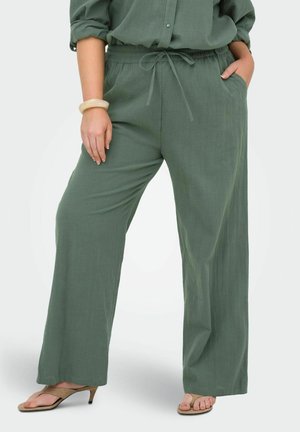 Kobieta w luźnych zielonych spodniach ze ściągaczem i kieszeniami oraz beżowych sandałkach na obcasie, stoi z jedną ręką w kieszeni, drugą wzdłuż ciała.