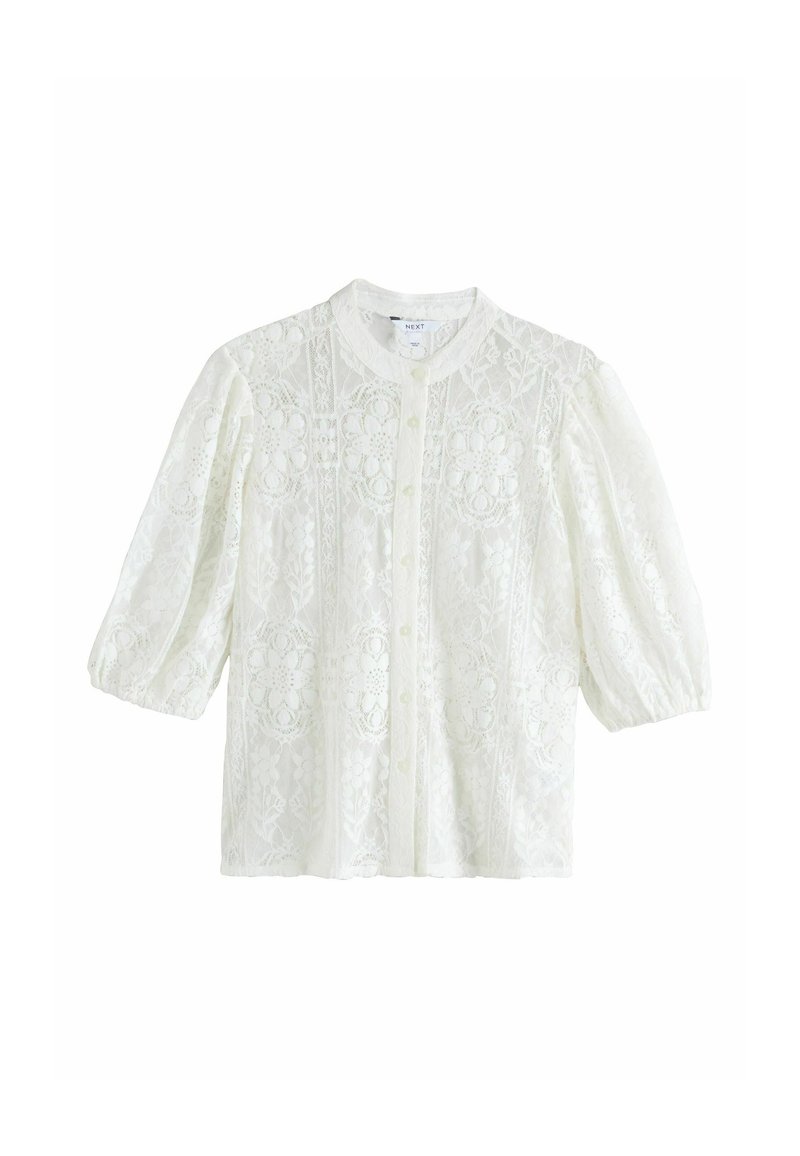 Balta mežģīņu blūze ar pūkainām piedurknēm, apaļu kaklu un dekoratīviem ziedu rakstiem. Ir pogājams priekšpuse un teksturēts audums.