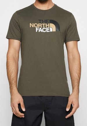 Mężczyzna noszący oliwkowo-zielony T-shirt z krótkim rękawem z logo "THE NORTH FACE" w kolorach czarnym, beżowym i białym na przodzie, w połączeniu z czarnymi spodniami.