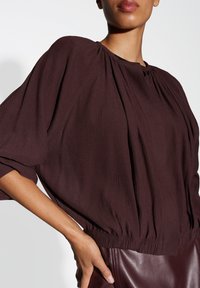 Bordeaux, textured blouse met een geraffineerde taille, losse ballonmouwen en een ronde halslijn. Heeft subtiele plooien voor extra detail.