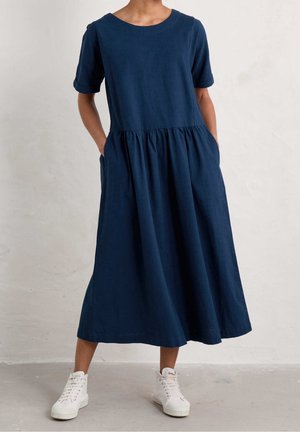Kobieta w luźnej granatowej sukience midi z krótkimi rękawami i bocznymi kieszeniami, w połączeniu z białymi wysokimi trampkami na jednolitym tle.