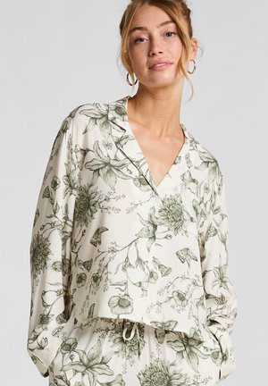 Woman wearing matching cream-colored floral-print top and pants with long sleeves and a drawstring waist, standing against a plain background.