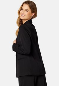 Zwarte blazer met een getailleerd ontwerp, sluiting met één knoop, scherpe revers en een subtiele textuur. Voorzien van twee zijzakken en een rugventilatie.