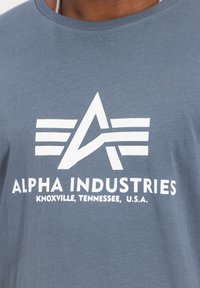 Graues Baumwoll-T-Shirt mit einem weißen Logo mit einem "A" und drei Streifen, darunter steht der Text "ALPHA INDUSTRIES KNOXVILLE, TENNESSEE, U.S.A."