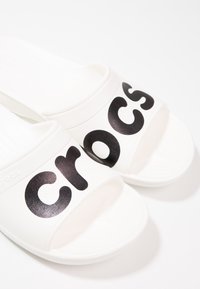 Sandales blanches à enfiler avec le texte noir "Crocs" sur les brides, sur un fond blanc uni.