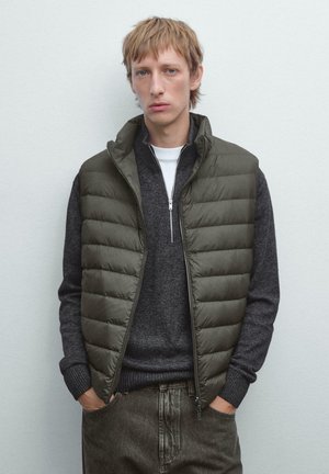 Giovane uomo che indossa un gilet imbottito oliva sopra un maglione grigio scuro a mezza zip, in piedi contro uno sfondo semplice grigio chiaro.