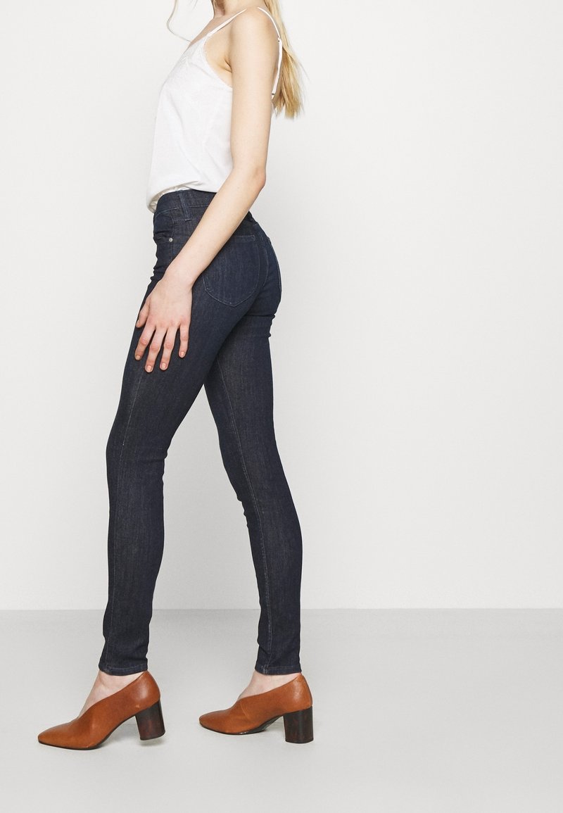 Smala mörka denimjeans tillsammans med en vit linne och bruna klackskor. Jeansen har en midja i mellanläge och en åtsittande silhuett.