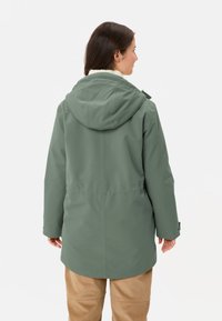 Giacca impermeabile verde con cappuccio e rivestimento in pelliccia sintetica, caratterizzata da una vestibilità dritta e polsini elasticizzati. Indossata sopra pantaloni beige.