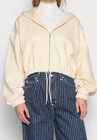 Krémový zkrácený svetr s kapucí na zip, se stahovací šňůrkou na spodním lemu a př oversized rukávy, v kombinaci s modrými džíny s vertikálními pruhy.