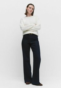 Vrouw met kort bruin haar staat zelfverzekerd en draagt een witte trui, donkere hoge taille wijde broek, bruine schoenen en een bruine riem.