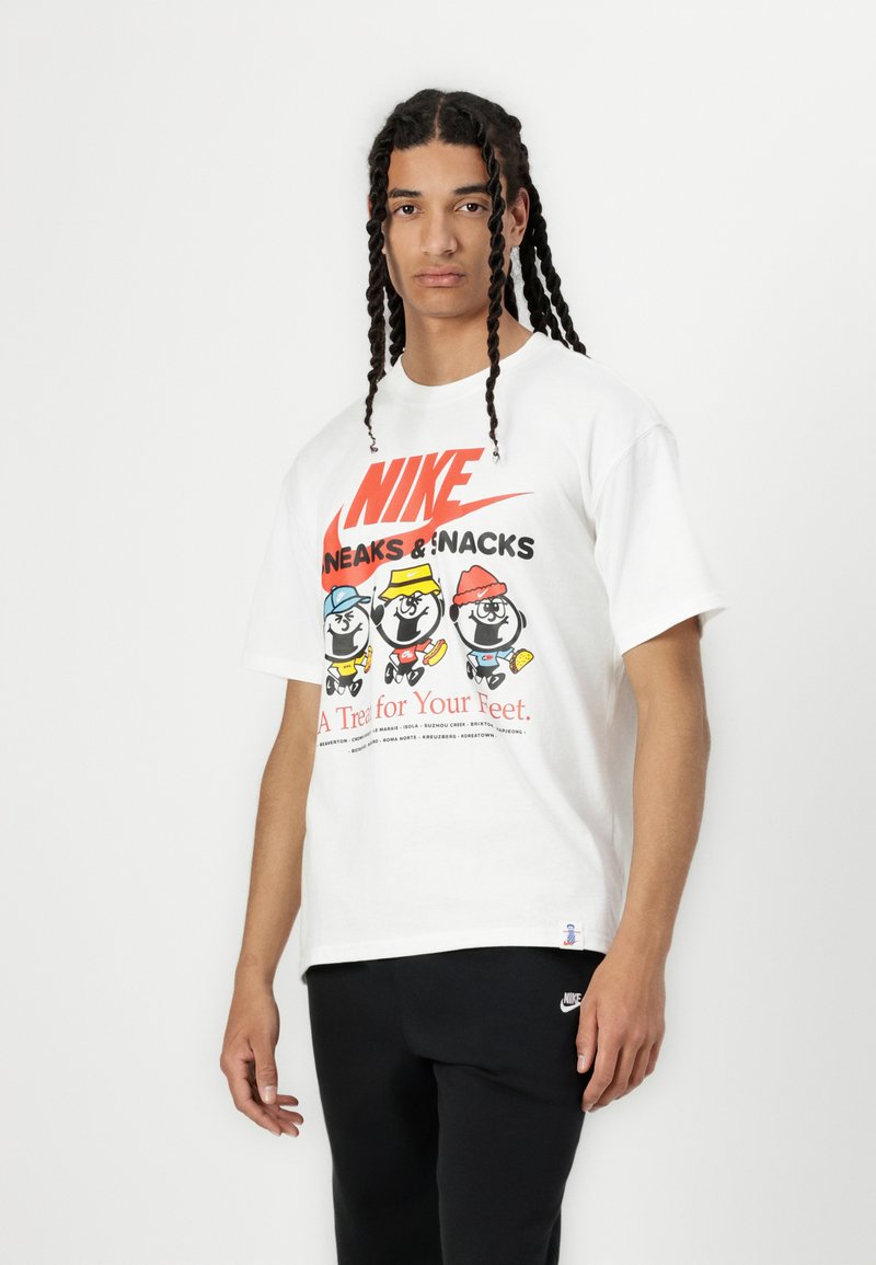 Bela bombažna majica z barvitimi risanimi junaki in besedilom, z rdečim logotipom Nike. V kombinaciji z črnimi hlačami in diskretnim logotipom.