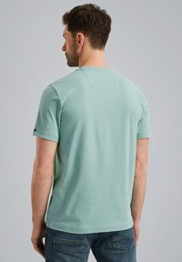 Camisa de algodón color verde menta con mangas cortas, cuello redondo y un parche rectangular en la parte posterior. Combinada con jeans de color azul oscuro.