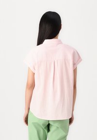 Camisa de mangas curtas em rosa e branco com riscas, com colarinho, corte descontraído e detalhe de prega nas costas, combinada com calças verdes.