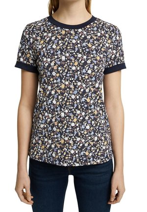 Kvinde iført en kortærmet marineblå skjorte med et flerfarvet lille blomstermønster og mørkeblå jeans.