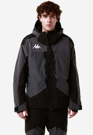 Giacca zip-up grigia e nera con cappuccio, caratterizzata da un tessuto testurizzato, logo sul petto e un prominente design a più pannelli. Maniche e zip decorate.