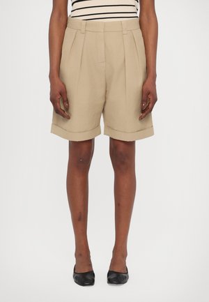 Beige høyhalsede shortser med folder, oppbrettede ermer og en skreddersydd design. Kombinert med svarte leiligheter og en stripete topp.