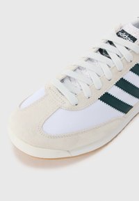 White sneaker with grey suede accents, green three-stripe logo, textured fabric panels, and a rubber sole. Metallic branding on tongue.