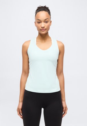 Jauna sieviete ar cirtainiem matiņiem sasietiem kūkā, valkājot gaiši zilu piedurknēm bez augšu un melnus legingus, stāvot pret gaiši pelēku fona. 