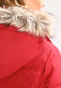 Femme portant un manteau d'hiver rouge avec une capuche ornée d'une bordure en fausse fourrure marron clair, vue de dos et de profil.