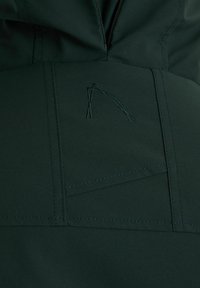 Temno zelena bluzon tkanina z diskretnim vezenjem logotipa v svetlejši zeleni barvi. Gladka tekstura z vidnimi šivi in podrobnostmi šivanja.