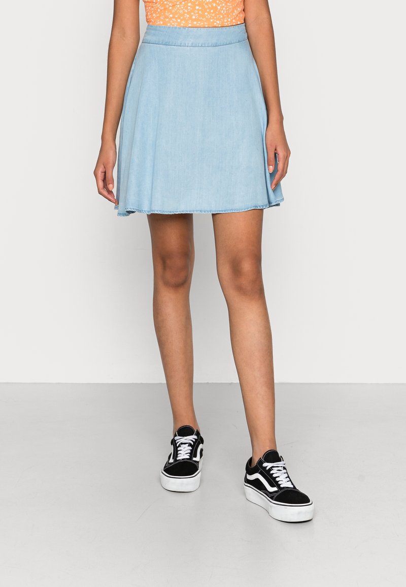 Hellblauer Denimrock mit ausgestelltem Design, ausgefranstem Saum und tailliertem Bund, kombiniert mit schwarzen und weißen Turnschuhen. Glatte Textur.