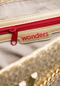 Tissu doré avec une finition texturée, fermeture éclair rouge et une étiquette portant l'inscription "merveilles" en rouge sur un fond beige.