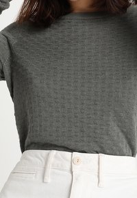 Pull en maille grise avec un col rond, présentant un motif en relief discret. Associé à un pantalon taille haute de couleur claire avec un détail de bouton.