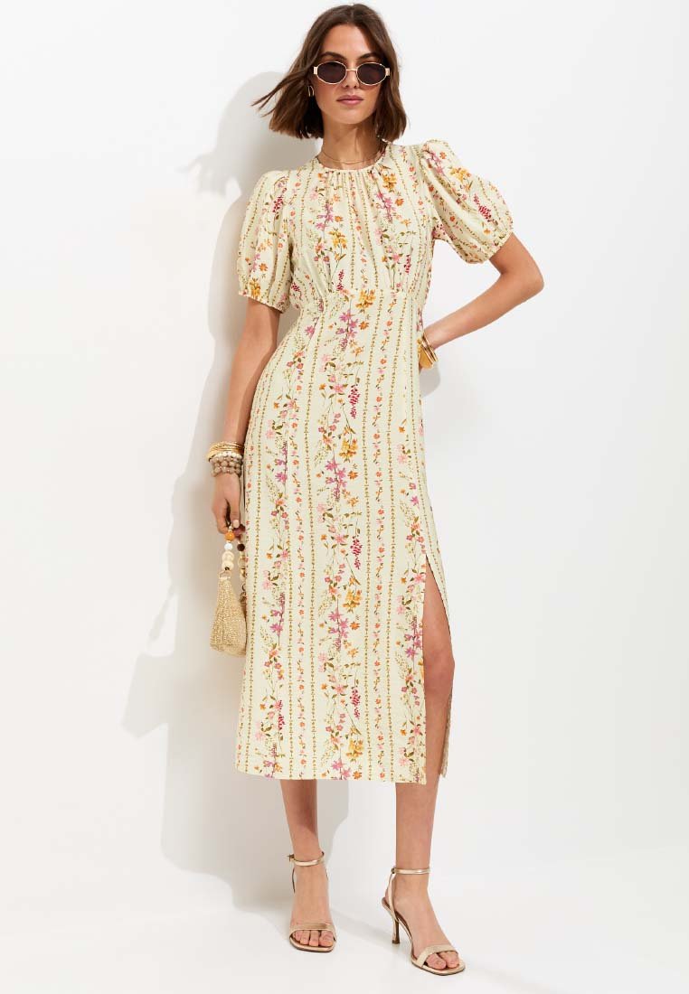 Vrouw met zonnebril draagt een lichte, bloemen midi-jurk met pofmouwen, splitrok, sandalen met hak, armbanden en houdt een kralentas vast.