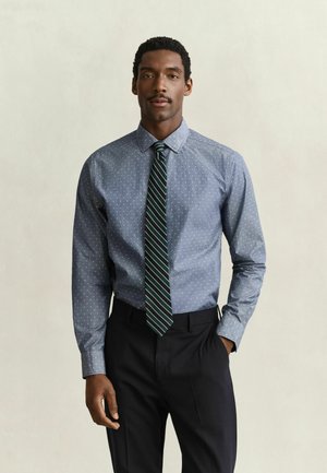 Uomo che indossa una camicia blu con piccoli puntini bianchi, una cravatta verde e bianca a righe e pantaloni neri, in piedi davanti a uno sfondo semplice.