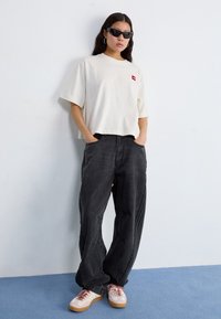 Ung kvinna som bär en vit oversize-t-shirt, svarta vida jeans, solglasögon och beige sneakers, stående mot en vit vägg på ett blått golv.