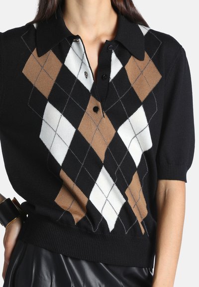Femme portant un pull noir à manches courtes avec un motif losanges marron, blanc et noir et une jupe en cuir noire, avec un bracelet noir au poignet.