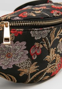 Černá látková ledvinka s barevným květinovým vyšíváním v červené, fialové a zlaté barvě. Disponuje zlatým zipem a kulatým tvarem.