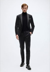 Schwarze Samtsakko mit Satinrevers, kombiniert mit einem eng anliegenden schwarzen Rollkragenpullover und passenden, maßgeschneiderten Hosen, vollendet mit schwarzen Schuhen.