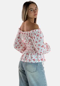 Witte off-shoulder blouse met roze bloemenpatroon, een gerimpeld lijfje en lange, opgeblazen mouwen. Gecombineerd met een hoge taille blauwe jeans.