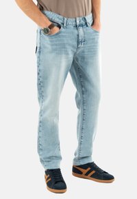 Jeans de mezclilla azul claro con un corte relajado, que cuentan con cinco bolsillos y una textura desgastada. Combinados con zapatillas negras y detalles en marrón.